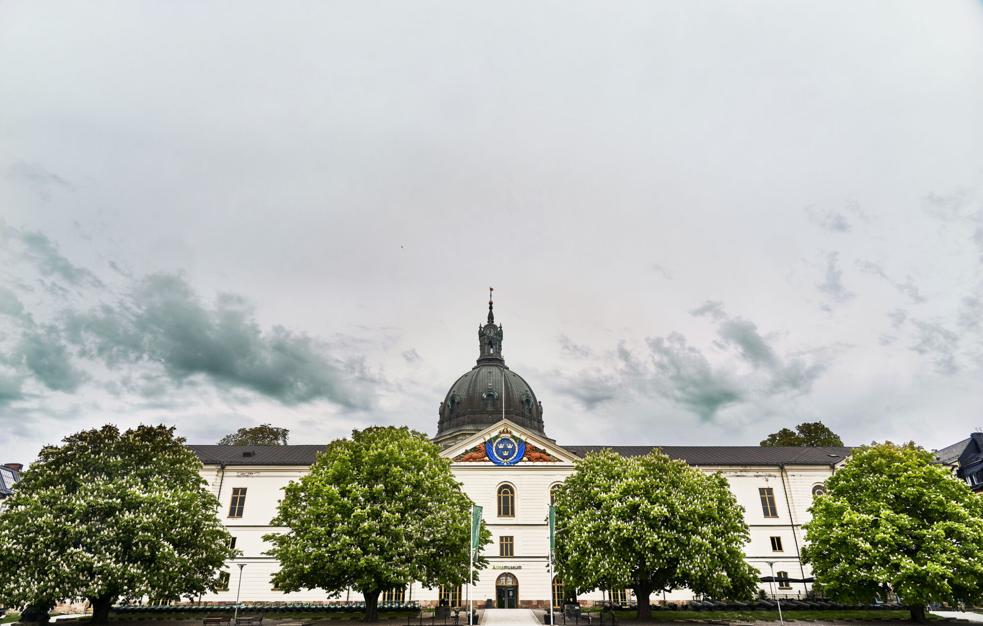 Vit byggnad med svart kupoltak &ouml;ver entr&eacute;n, sedd fr&aring;n gatan omgiven av gr&ouml;na tr&auml;d under en molnig himmel.
