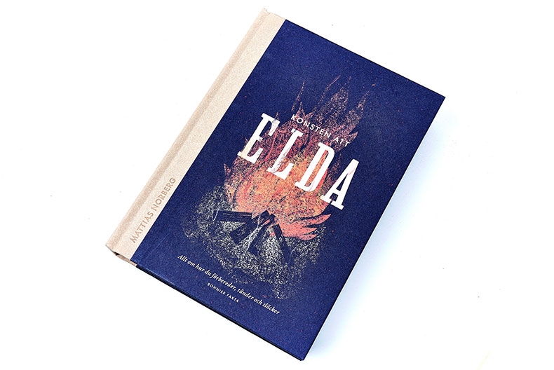 Bokomslad till boken "Elda", Vitt band och bl&aring; framsida med en r&ouml;d eld bakom bokens titel.