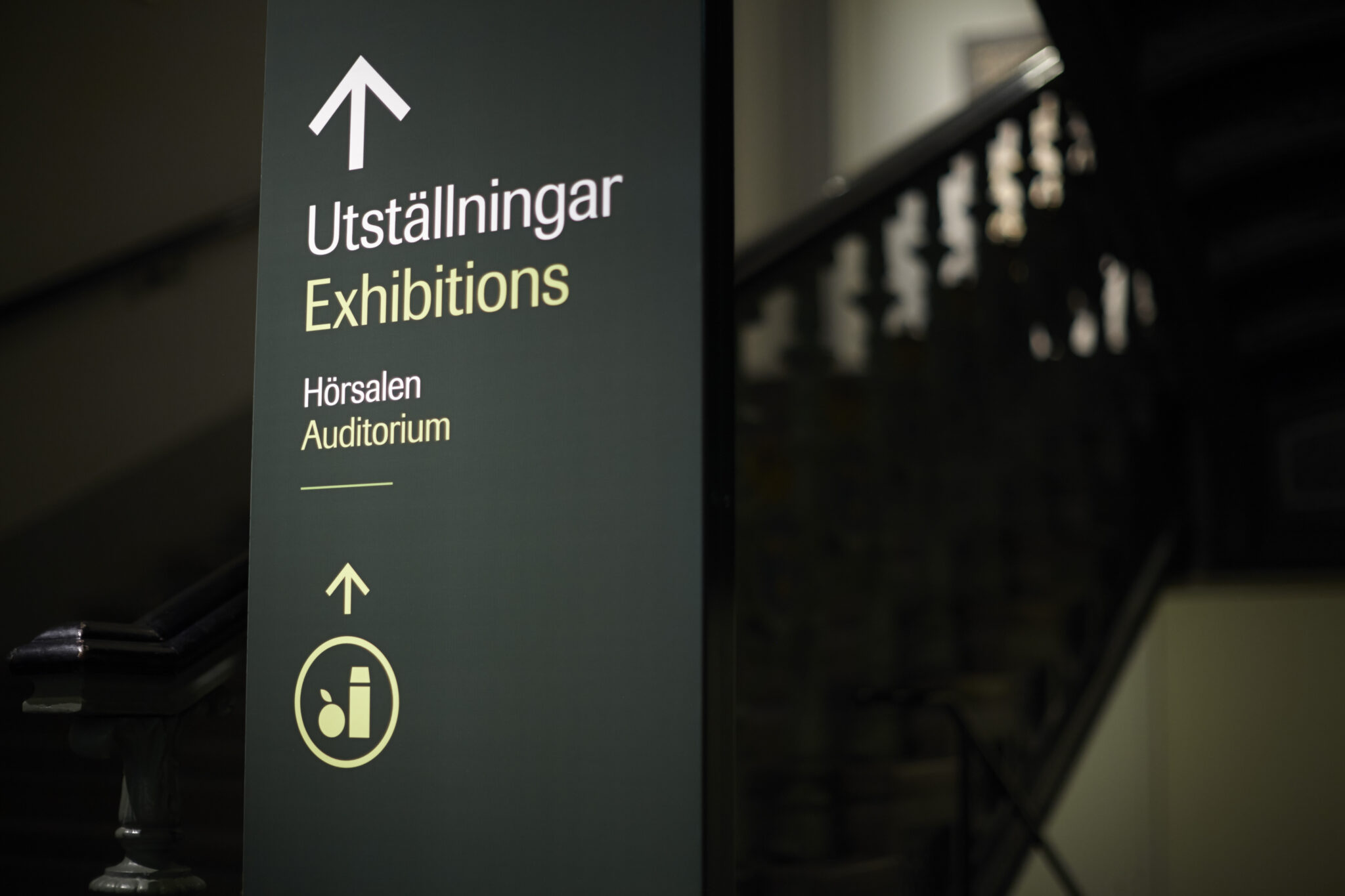 En gr&ouml;n skylt som det st&aring;r "Utst&auml;llningar" p&aring; samt en pil som pekar upp&aring;t. I bakgrunden syns en trappa upp till n&auml;sta v&aring;ning.
