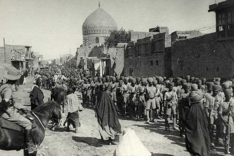 Svartvit bild av milit&auml;rparad p&aring; en stadsgata, med rader av soldater i turbaner och korta uniformer. I bakgrunden syns en byggnad med kupoltak.