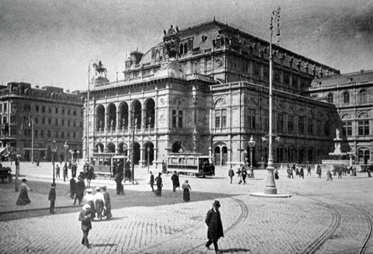 En stor pampig byggnad, Wiens statsopera. P&aring; det stenbelagda torget framf&ouml;r operan st&aring;r tv&aring; &auml;ldre sp&aring;rvagnar och m&auml;nniskor promenerar omkring.
