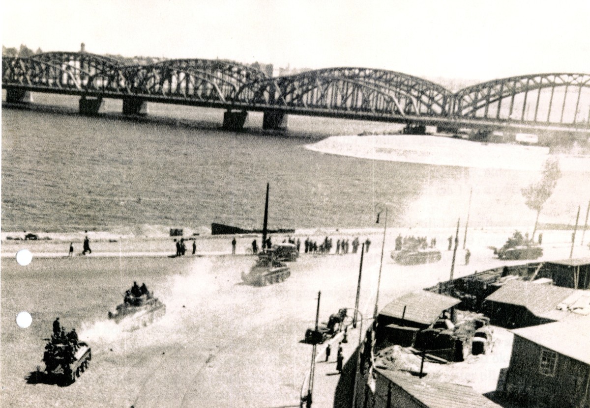 Milit&auml;rfordon k&ouml;r l&auml;ngs vatten. L&auml;ngre bort syns en l&aring;ng bro.