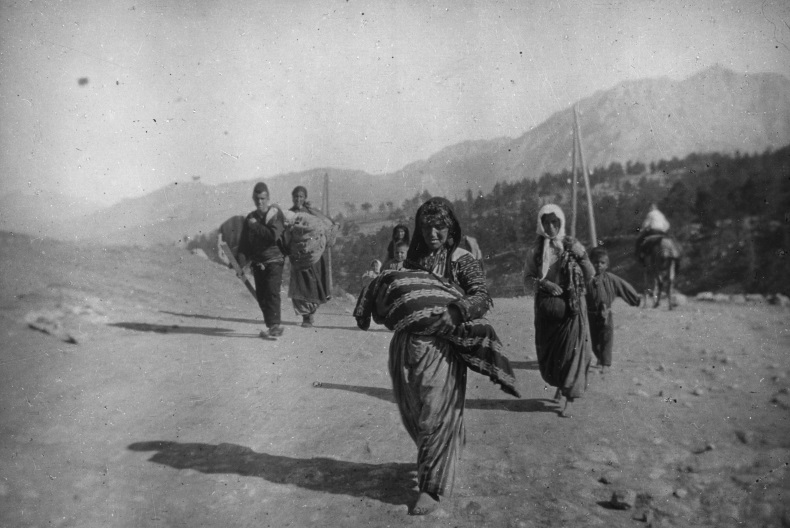 Svartvit bild av m&auml;nniskor p&aring; flykt som g&aring;r p&aring; en v&auml;g med skog och berg i bakgrunden. L&auml;ngst fram en kvinna med ett stort bylte i famnen.