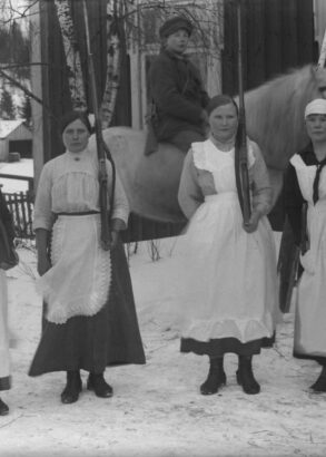 Framf&ouml;r ett hus st&aring;r fyra personer if&ouml;rda kl&auml;nning och vita f&ouml;rkl&auml;den p&aring; rad med varsitt gev&auml;r lutat mot v&auml;nster axel, bakom dem sitter en person till h&auml;st. Det &auml;r vinter.