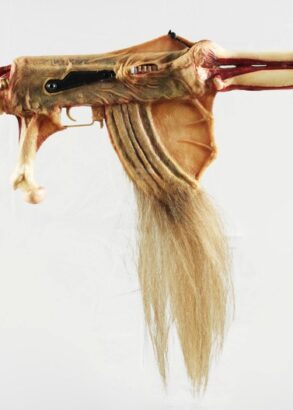 Ett konstn&auml;rligt vapenliknande f&ouml;rem&aring;l skapat av material som p&aring;minner om djurben, hud och h&aring;r. Objektet har formen av ett gev&auml;r men best&aring;r av organiska former med en ryggradsliknande kolv, benformade detaljer och en l&aring;ng tofs av ljust h&aring;r som h&auml;nger fr&aring;n undersidan. Bakgrunden &auml;r helt vit.