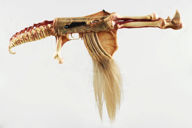 Ett konstn&auml;rligt vapenliknande f&ouml;rem&aring;l skapat av material som p&aring;minner om djurben, hud och h&aring;r. Objektet har formen av ett gev&auml;r men best&aring;r av organiska former med en ryggradsliknande kolv, benformade detaljer och en l&aring;ng tofs av ljust h&aring;r som h&auml;nger fr&aring;n undersidan. Bakgrunden &auml;r helt vit.