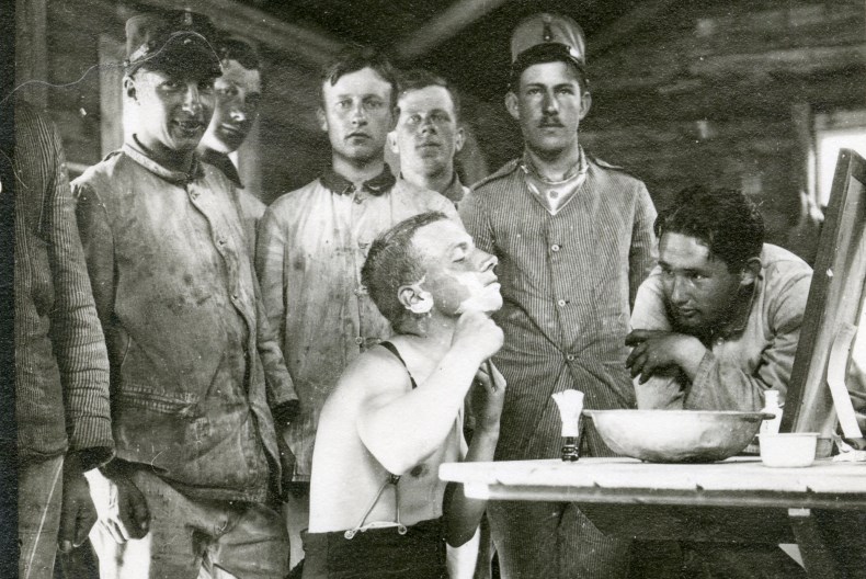 En person sitter framf&ouml;r spegeln vid ett bord med en vattensk&aring;l framf&ouml;r sig och rakar sig medan sju andra personer tittar p&aring;.