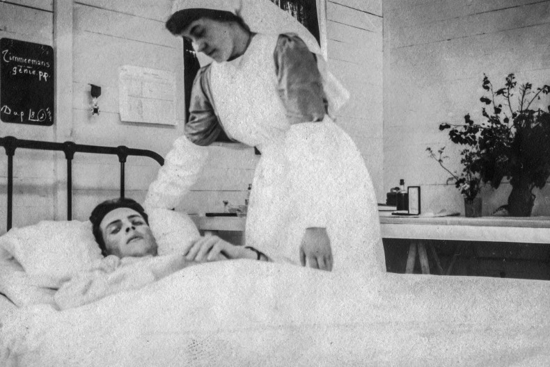 En sk&ouml;terska i sk&ouml;terskeuniform b&ouml;jer sig ner &ouml;ver en skadad soldat nedb&auml;ddad i en s&auml;ng med vita lakan.