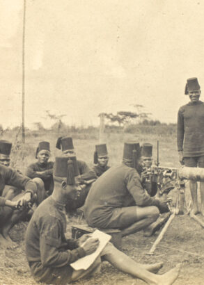 Svartvit bild av soldater i f&auml;lt. flera sittandes p&aring; marken och n&aring;gra st&aring;ende vid ett maskingev&auml;r med en officer i hj&auml;lm bredvid.