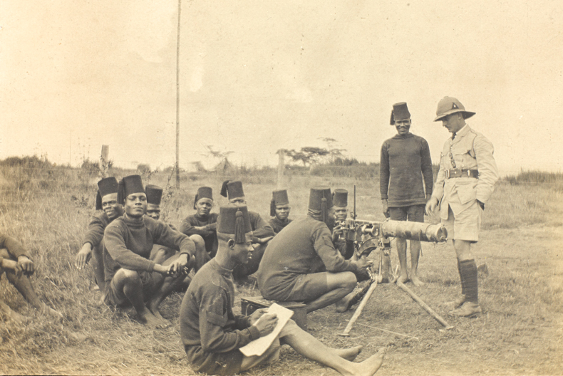 Svartvit bild av soldater i f&auml;lt. flera sittandes p&aring; marken och n&aring;gra st&aring;ende vid ett maskingev&auml;r med en officer i hj&auml;lm bredvid.