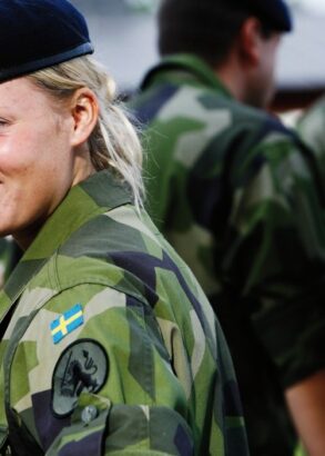 En soldat i kamouflagekl&auml;der med FN:s v&auml;lk&auml;nda bl&aring; basker, halvt v&auml;nd mot kameran. I bakgrunden fyra andra soldater.