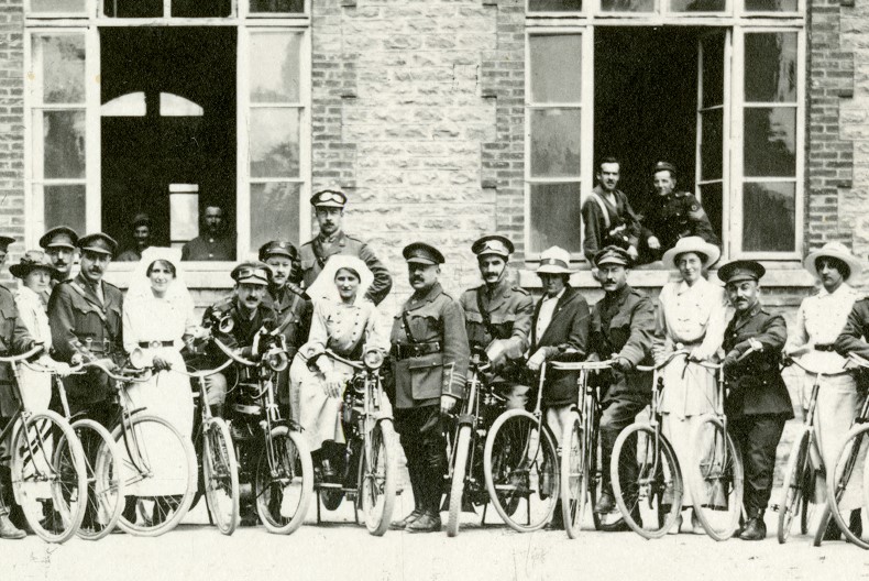 En grupp kvinnor och m&auml;n i uniform och sjukv&aring;rdkl&auml;der st&aring;r och poserar med cyklar utanf&ouml;r en tegelbyggnad med vid&ouml;ppna f&ouml;nster d&auml;r fler personer skymtas.