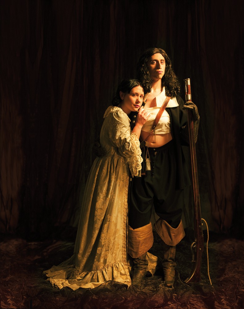 En person i vit b&ouml;ljande kl&auml;nning lutar sig mot en annan person st&ouml;dd p&aring; sitt gev&auml;r och kl&auml;dd i uniform. Uniformsjackan &auml;r &ouml;ppen och br&ouml;stet lindat med vitt tyg.