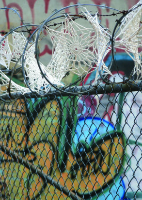 Framf&ouml;r en graffittim&aring;lad v&auml;gg st&aring;r ett staket med taggtr&aring;d. Taggtr&aring;den &auml;r dekorerad med virkade sm&aring;dukar som p&aring;minner om spindeln&auml;t d&auml;r de sitter.