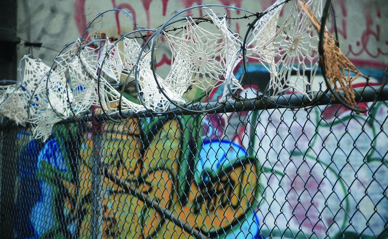 Framf&ouml;r en graffittim&aring;lad v&auml;gg st&aring;r ett staket med taggtr&aring;d. Taggtr&aring;den &auml;r dekorerad med virkade sm&aring;dukar som p&aring;minner om spindeln&auml;t d&auml;r de sitter.