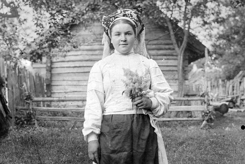 En person st&aring;r framf&ouml;r ett timrat hus med gr&ouml;nska runtom, hon har en huvudbonad, vit blus och troligtvis en kjol p&aring; sig. I v&auml;nstra handen h&aring;ller hon en blombukett.