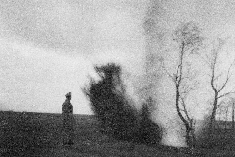 En milit&auml;rkl&auml;dd person med vapen i handen st&aring;r p&aring; ett f&auml;lt, med bara n&aring;got enstaka tr&auml;d, och ser bort i fj&auml;rran.