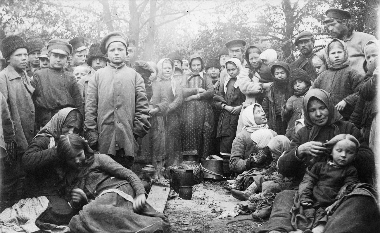 En stor grupp m&auml;nniskor, varav m&aring;nga barn, kl&auml;dd i ytterkl&auml;der och ofta med n&aring;got p&aring; huvudet, de flesta st&aring;r upp, n&aring;gra sitter ner p&aring; marken. De flesta tittar in i kameran. I mitten p&aring; marken kastruller och k&auml;rl.