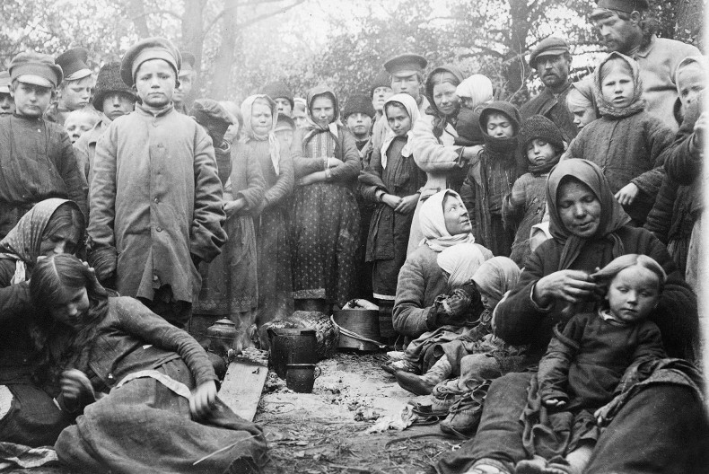 En stor grupp m&auml;nniskor, varav m&aring;nga barn, kl&auml;dd i ytterkl&auml;der och ofta med n&aring;got p&aring; huvudet, de flesta st&aring;r upp, n&aring;gra sitter ner p&aring; marken. De flesta tittar in i kameran. I mitten p&aring; marken kastruller och k&auml;rl.
