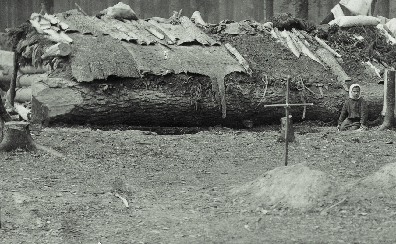 I f&ouml;rgrunden ett kors av pinnar nedstucket i jorden. I bakgrunden ett barn som sitter framf&ouml;r ett f&auml;llt tr&auml;d som utg&ouml;r basen i en stuga med lappat tak.