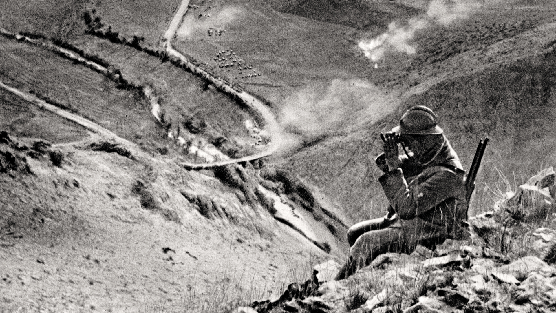 En man med vapen p&aring; ryggen sitter p&aring; en h&ouml;jd i bergen och spanar med sin kikare. L&auml;ngre bort syns en v&auml;g som snirklar sig upp&aring;t.