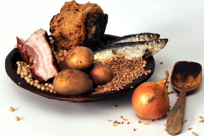 En pl&aring;ttallrik med en bit br&ouml;d, tre oskalade potatisar, tv&aring; hela fiskar, en bit fl&auml;sk med sv&aring;len kvar och spannm&aring;l. Bredvid en oskalad gul l&ouml;k och en tr&auml;sked.