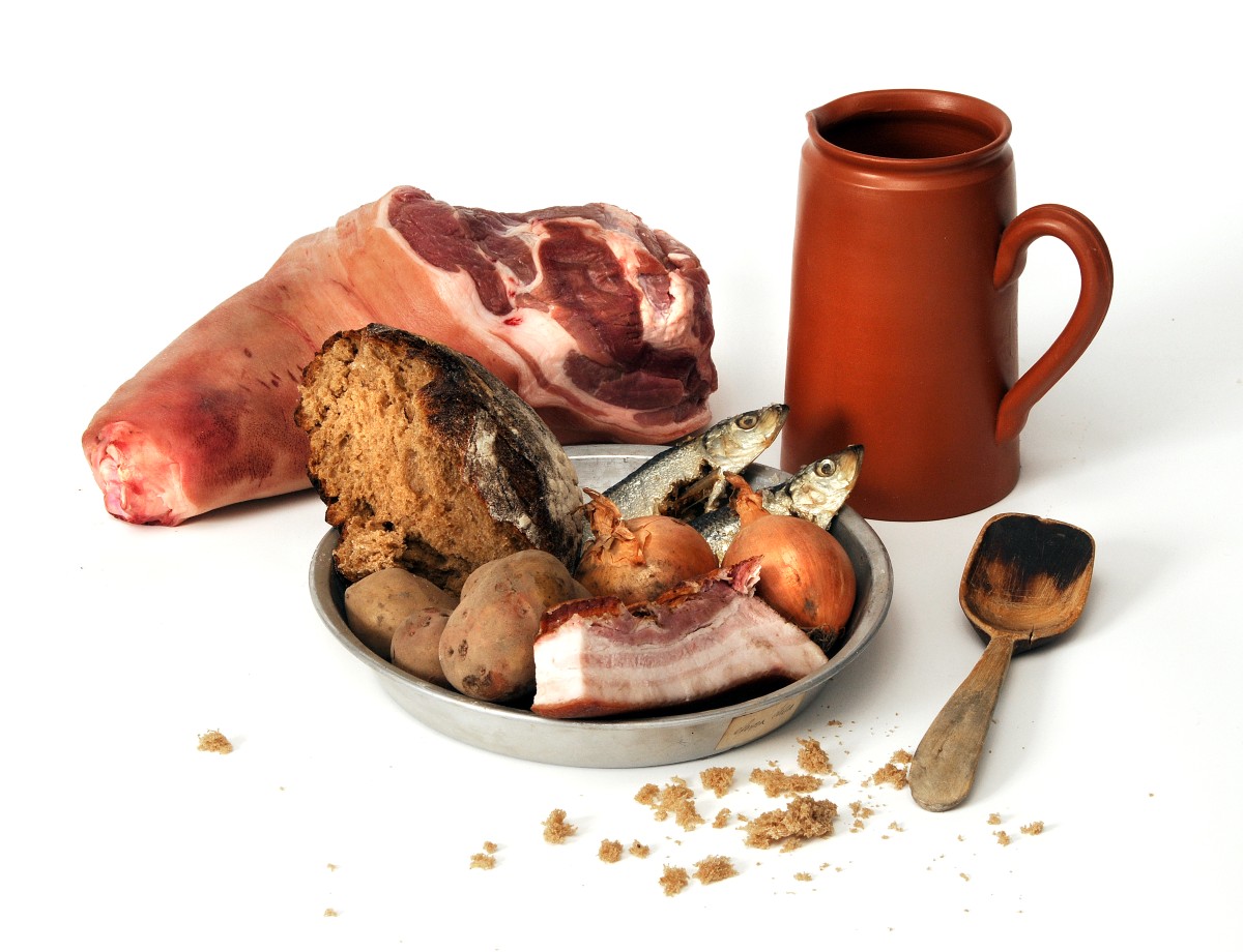 En pl&aring;ttallrik med en bit br&ouml;d, fyra oskalade potatisar, tv&aring; hela fiskar, tv&aring; oskalade gull&ouml;kar och bit fl&auml;sk med sv&aring;len kvar. Bredvid en tr&auml;sked och bakom tallriken ytterligare en bit k&ouml;tt samt ett lerkrus.