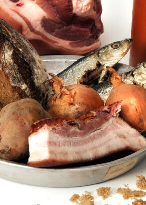 En pl&aring;ttallrik med en bit br&ouml;d, fyra oskalade potatisar, tv&aring; hela fiskar, tv&aring; oskalade gull&ouml;kar och bit fl&auml;sk med sv&aring;len kvar. Bredvid en tr&auml;sked och bakom tallriken ytterligare en bit k&ouml;tt samt ett lerkrus.