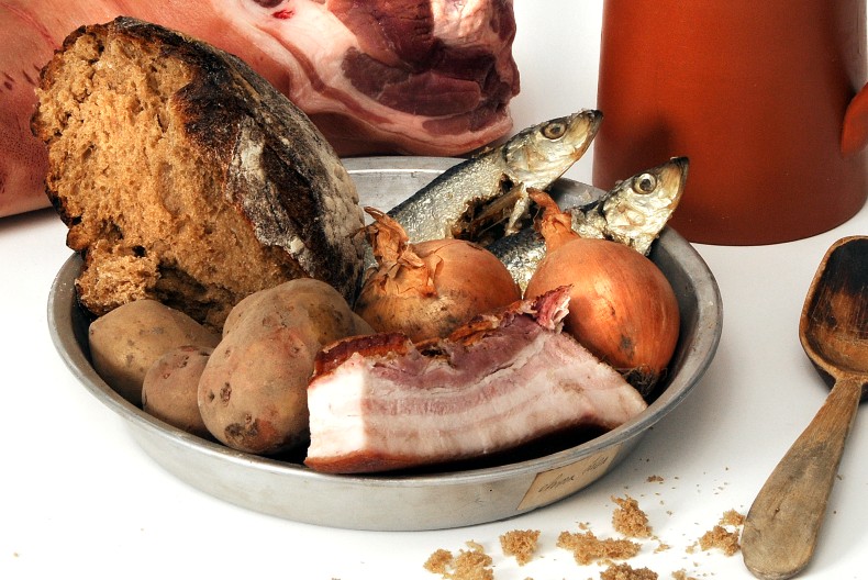 En pl&aring;ttallrik med en bit br&ouml;d, fyra oskalade potatisar, tv&aring; hela fiskar, tv&aring; oskalade gull&ouml;kar och bit fl&auml;sk med sv&aring;len kvar. Bredvid en tr&auml;sked och bakom tallriken ytterligare en bit k&ouml;tt samt ett lerkrus.