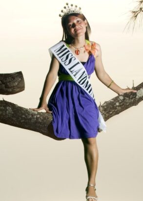 En person i lila kl&auml;nning, tiara i h&aring;ret och med ett satinband &ouml;ver br&ouml;stet "Miss landmine" sitter p&aring; en tr&auml;dgren.