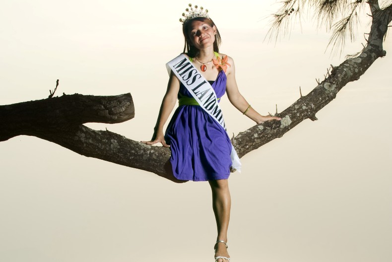 En person i lila kl&auml;nning, tiara i h&aring;ret och med ett satinband &ouml;ver br&ouml;stet "Miss landmine" sitter p&aring; en tr&auml;dgren.