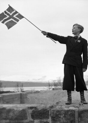 En person st&aring;r p&aring; en stenmur och h&aring;ller stolt upp Norges flagga.