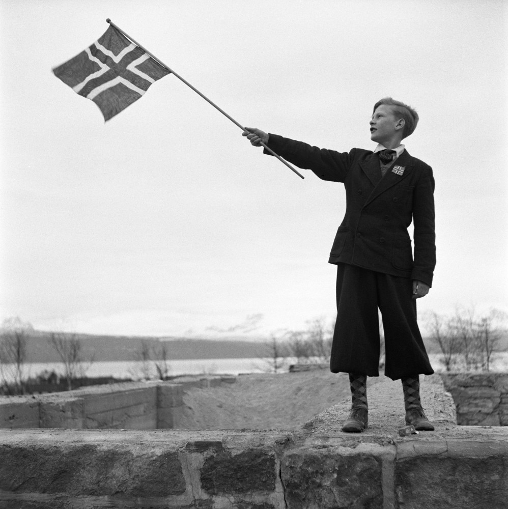 En person st&aring;r p&aring; en stenmur och h&aring;ller stolt upp Norges flagga.