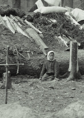 I f&ouml;rgrunden ett kors av pinnar nedstucket i jorden. I bakgrunden ett barn som sitter framf&ouml;r ett f&auml;llt tr&auml;d som utg&ouml;r basen i en stuga med lappat tak.