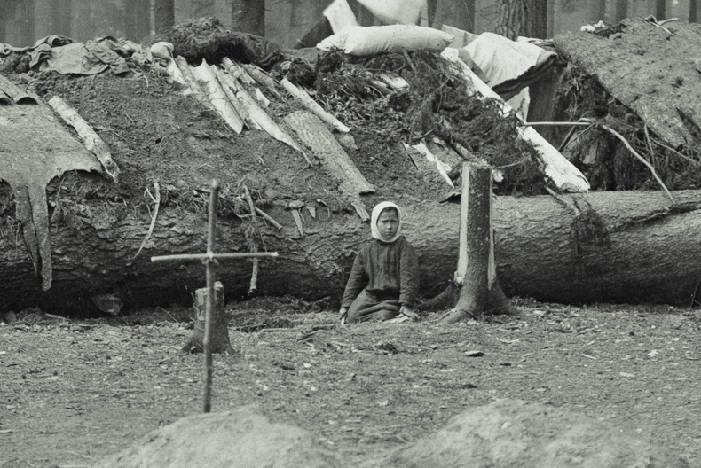 I f&ouml;rgrunden ett kors av pinnar nedstucket i jorden. I bakgrunden ett barn som sitter framf&ouml;r ett f&auml;llt tr&auml;d som utg&ouml;r basen i en stuga med lappat tak.
