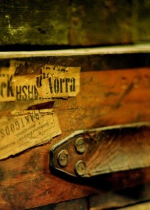 En n&auml;rbild av en gammal koffert i tr&auml; med etiketter p&aring;, d&auml;r handtaget i l&auml;der har g&aring;tt av.