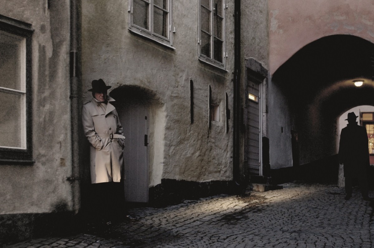 Tv&aring; personer i en m&ouml;rk gr&auml;nd. Ikl&auml;dda trenchcoats och hattar.