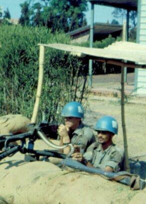 Tv&aring; soldater med bl&aring; hj&auml;lmar tittar upp bakom en mur av bruna sands&auml;ckar. Bredvid dem &auml;r det buskage och bakom dem ett tr&auml;hus.
