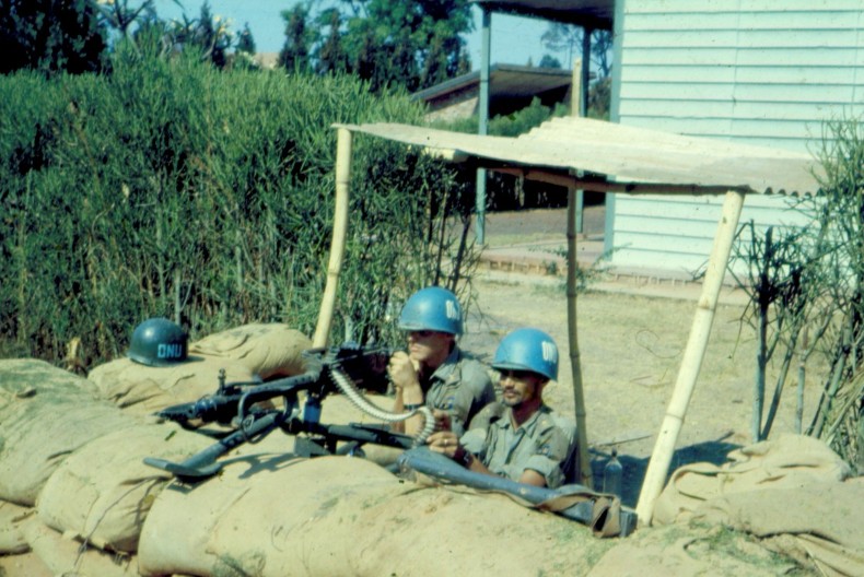 Tv&aring; soldater med bl&aring; hj&auml;lmar tittar upp bakom en mur av bruna sands&auml;ckar. Bredvid dem &auml;r det buskage och bakom dem ett tr&auml;hus.