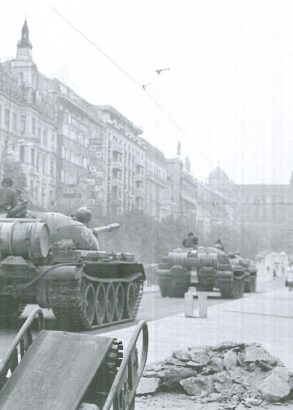 Tv&aring; stridsvagnar k&ouml;r genom en stad, en person i rock och med portf&ouml;lj st&aring;r bredvid och tittar p&aring;.