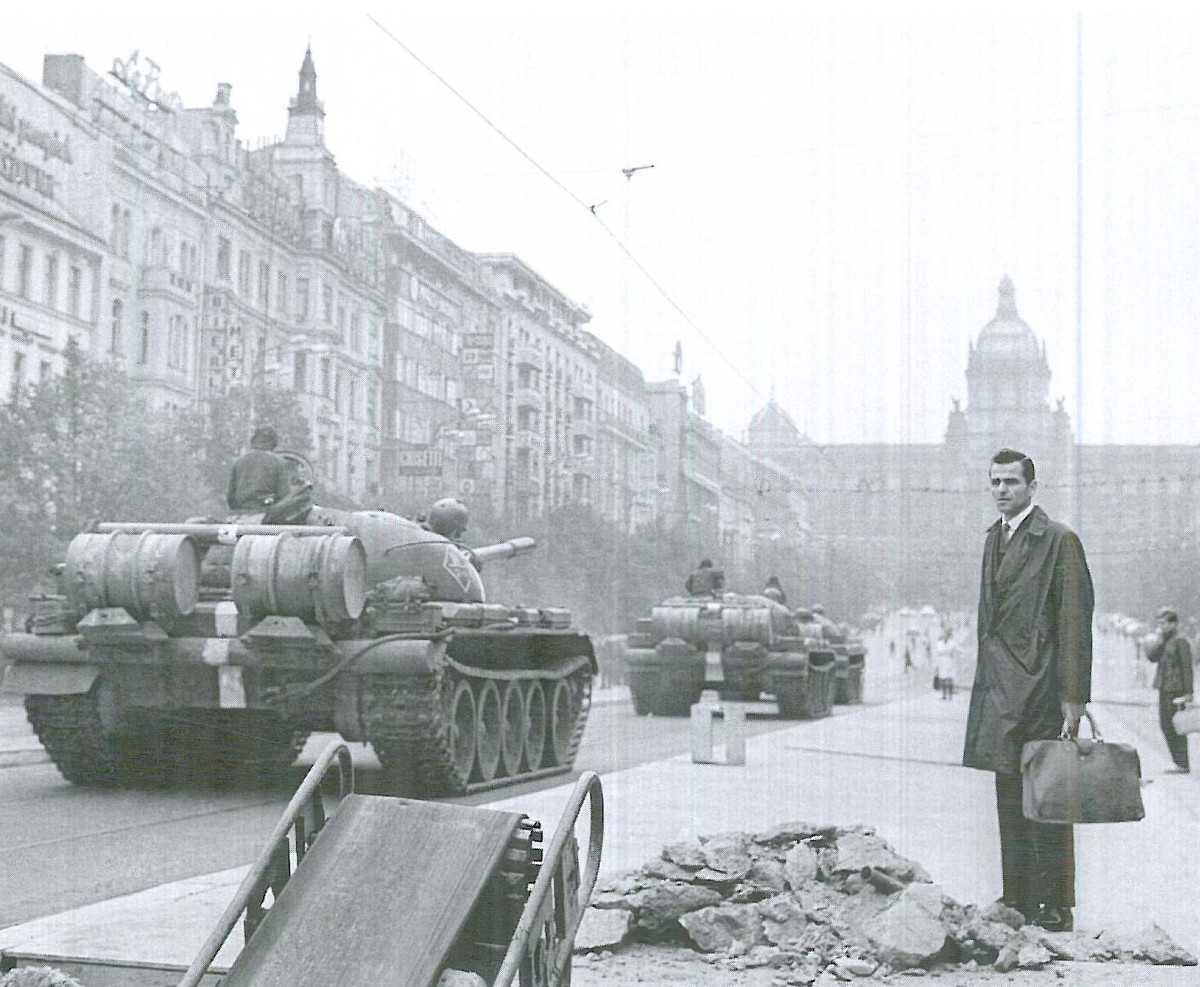 Tv&aring; stridsvagnar k&ouml;r genom en stad, en person i rock och med portf&ouml;lj st&aring;r bredvid och tittar p&aring;.