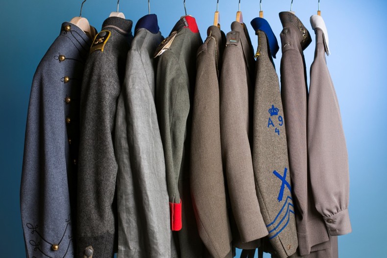 Nio uniformsjackor h&auml;nger p&aring; galgar p&aring; rad. Sm&aring; detaljer skiljer dem &aring;t s&aring;som olika gr&aring; nyanser, ett r&ouml;tt slag, bl&aring; rev&auml;rer.
