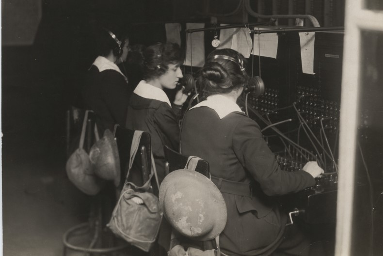 Tre telefonister med h&ouml;rlurar p&aring; sitter med ryggarna mot fotografen. Framf&ouml;r dem v&auml;xlingsbord med sladdar och knappar. P&aring; stolarna har de h&auml;ngt sina v&auml;skor.