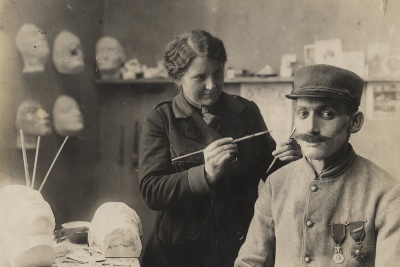 En del av ett rum, p&aring; v&auml;ggarna sitter gipsansikten/huvuden och p&aring; en hylla finns ytterligare n&aring;got huvud. En person i uniform och utm&auml;rkelser p&aring; br&ouml;stet sitter ner och verkar fungera som modell f&ouml;r personen som st&aring;r upp framf&ouml;r ett bord med ytterliga gipshuvuden.