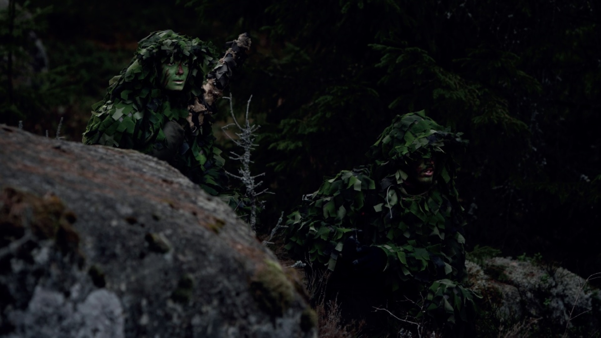 Kamouflagekl&auml;dda och gr&ouml;nm&aring;lade i ansiktena g&ouml;mmer sig n&aring;gra milit&auml;rer i skogen och spejar ut ur bild.