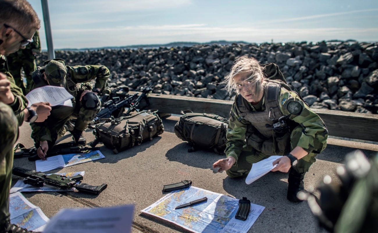 Tre kamouflagekl&auml;dda milit&auml;rer sitter p&aring; huk och studerar kartor. Omkring dem ligger deras packning samt vapen. Bakom dem en massa st&ouml;rre stenar och havet som skymtas.