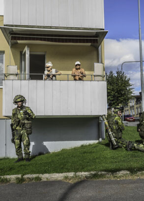 Ett &auml;ldre par st&aring;r p&aring; en balkong i ett hyreshus, nedanf&ouml;r dem v&auml;ntar fyra kamouflagekl&auml;dda personer med vapen.