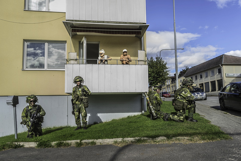 Ett &auml;ldre par st&aring;r p&aring; en balkong i ett hyreshus, nedanf&ouml;r dem v&auml;ntar fyra kamouflagekl&auml;dda personer med vapen.