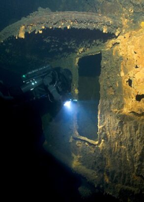 Ett del av ett vrak under vatten och en dykare som lyser med en lampa in i en &ouml;ppning.