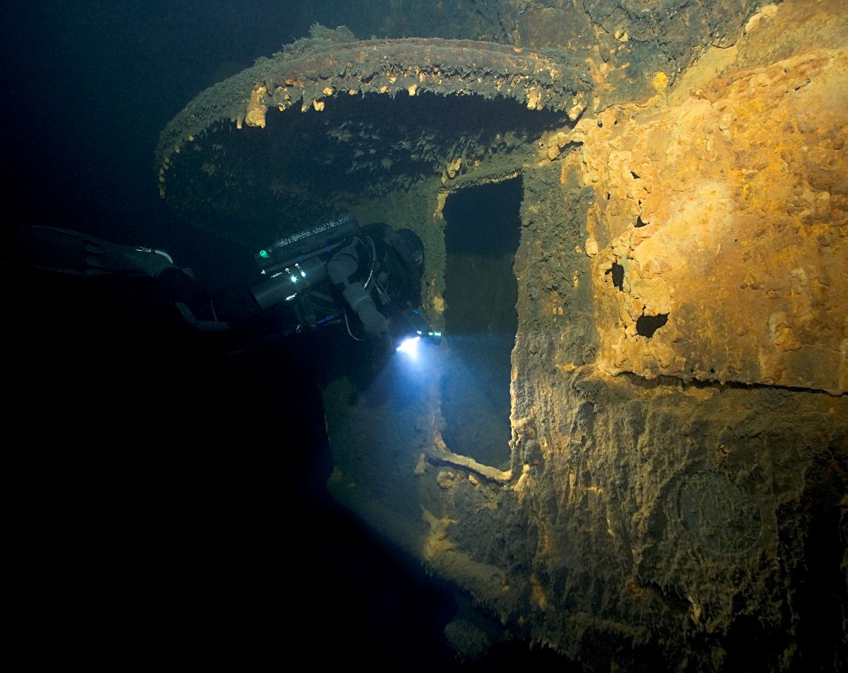 Ett del av ett vrak under vatten och en dykare som lyser med en lampa in i en &ouml;ppning.